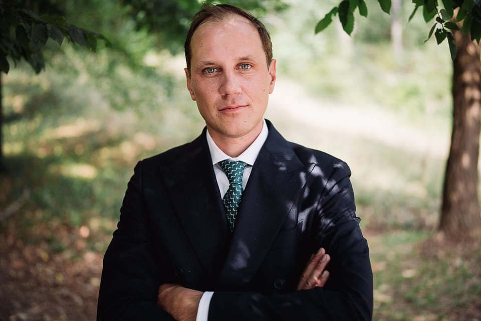 Egor Gorshkov in a dark suit and green tie, arms crossed, standing under trees.
